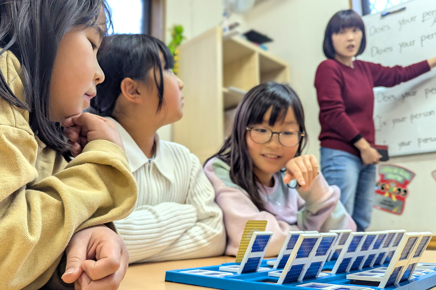 Elementary school students playing an English learning game while the teacher explains in the background
